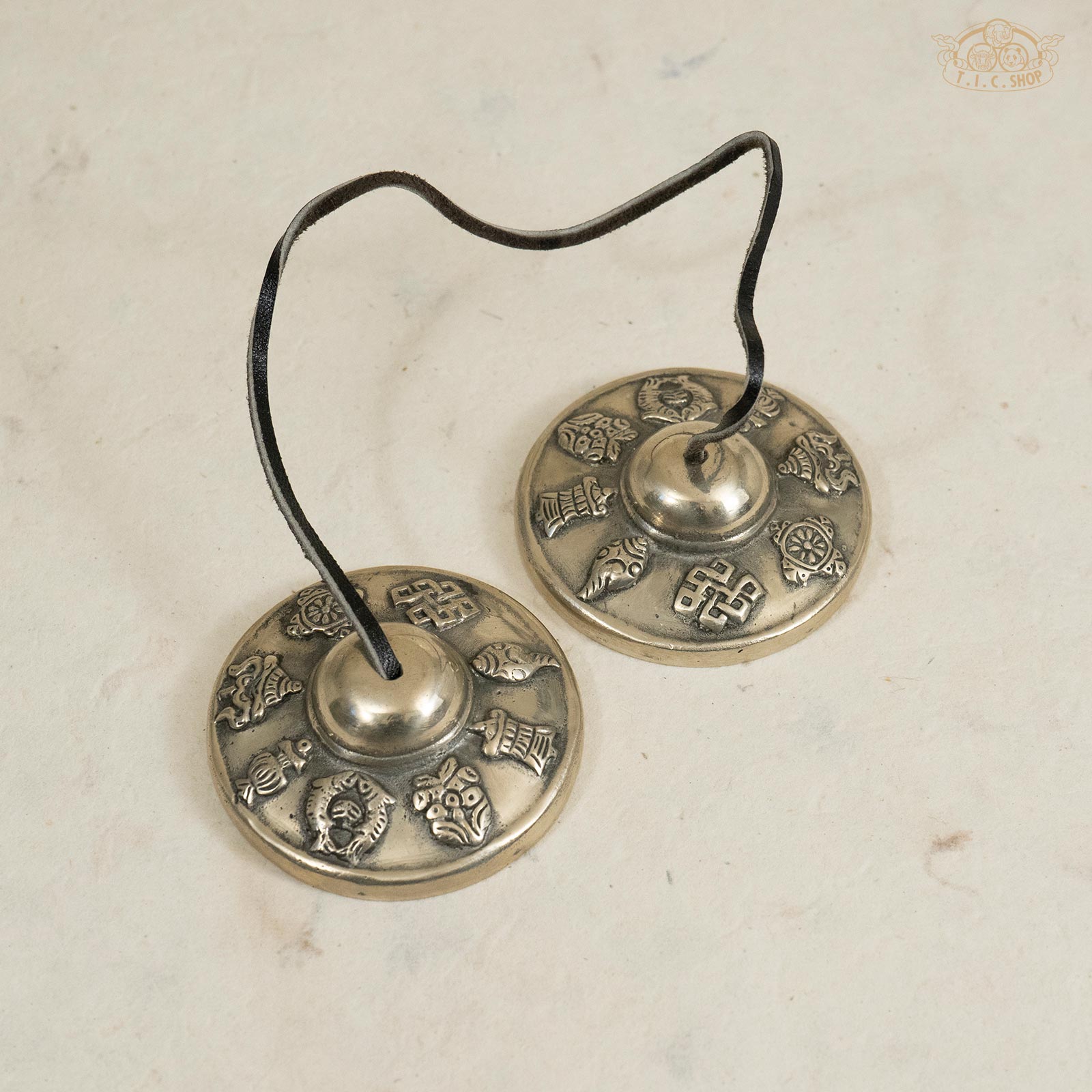 Front view of Tibetan handmade brass Tingsha bells decorated with the 8 auspicious symbols and connected by a leather strap.