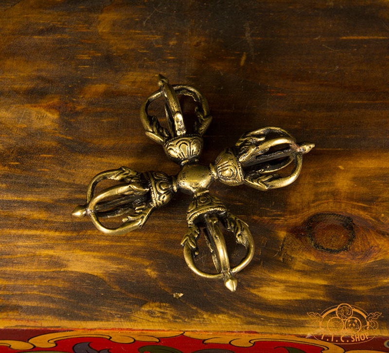 A top-down view of a handmade five-pronged brass Double Vajra (Vishvavajra) ritual implement, featuring intricate traditional carvings and a warm antique finish on a wooden surface.