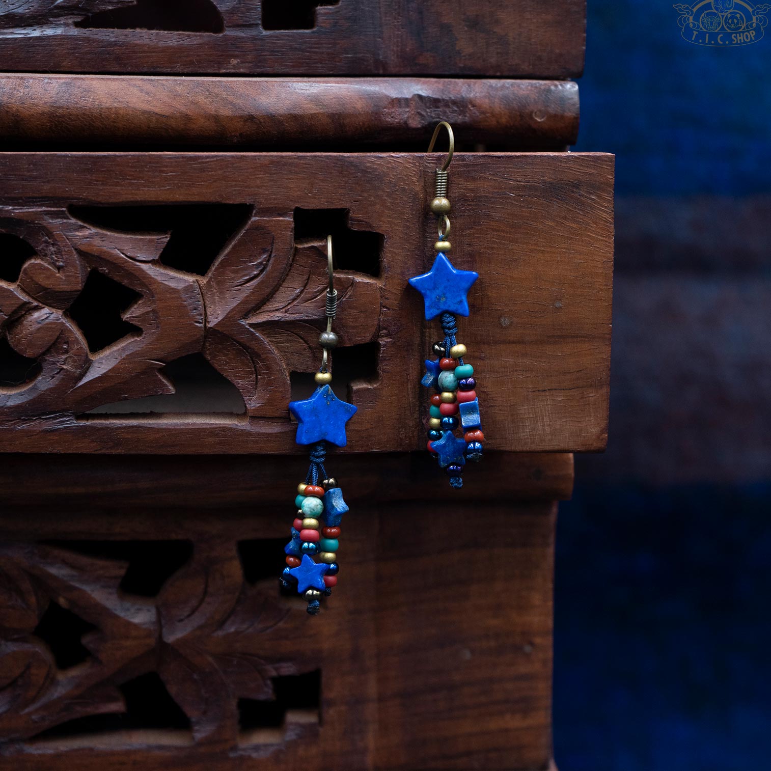 A pair of colorful handmade earrings with lapis lazuli stars, gold hooks, and multicolored beads, displayed hanging on a wooden surface.