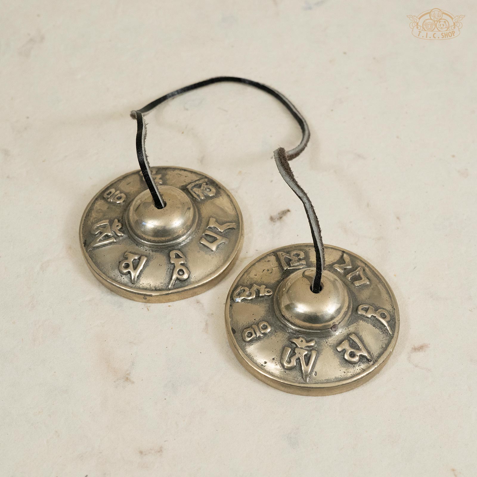 Front view of Tibetan handmade brass Tingsha bells engraved with the six-syllable mantra Om Mani Padme Hum, connected by a leather strap.